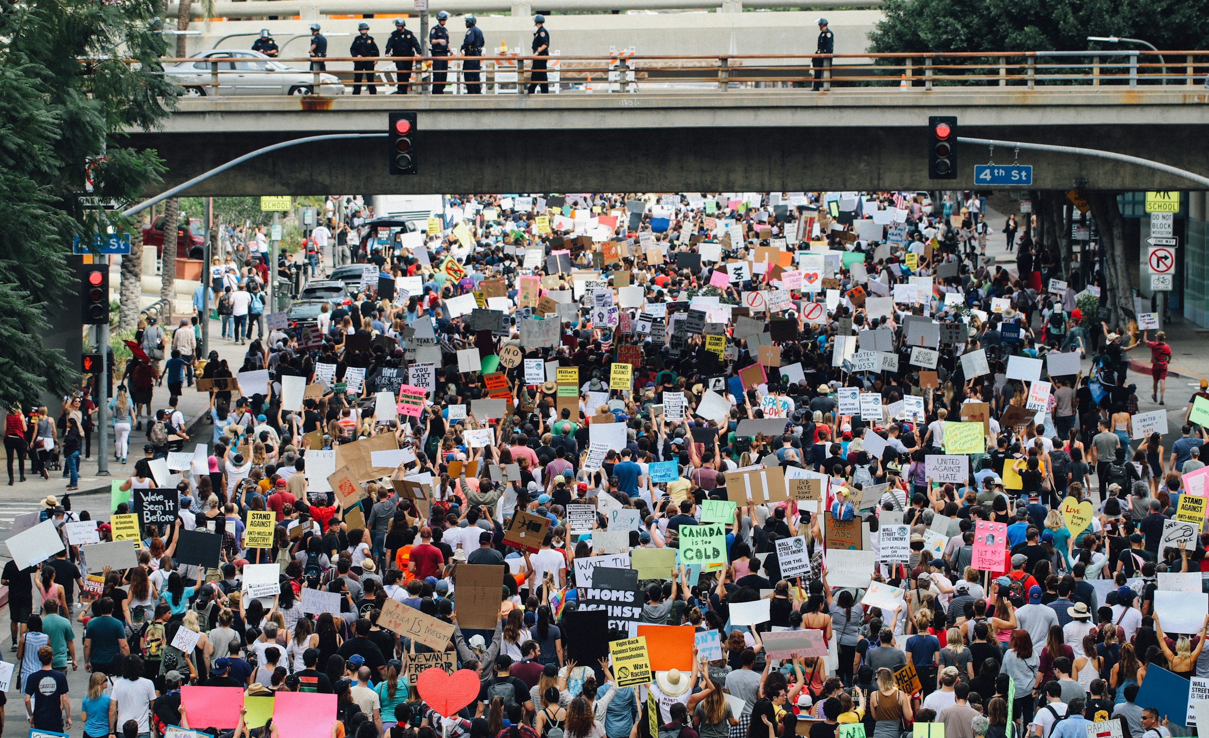 A large group of protesters holding up signs on the streets of Los Angeles, with police officers watching from a bridge above the crowd.