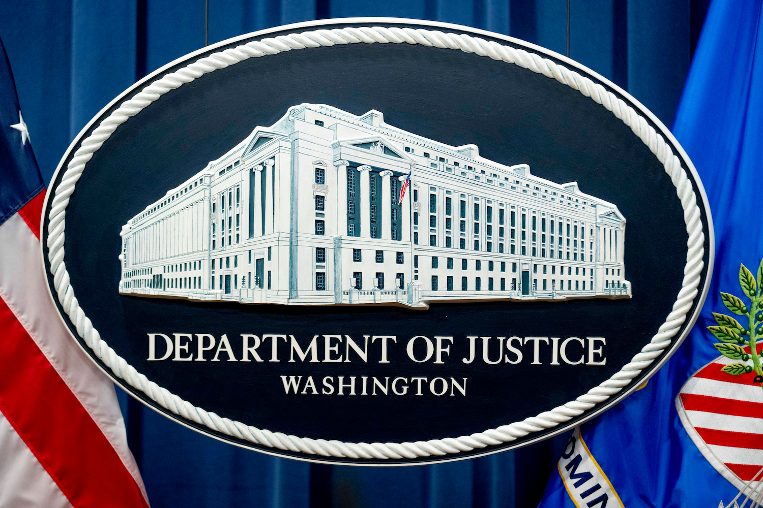 A sign depicting the Justice Department building hangs in front of dark blue curtains.